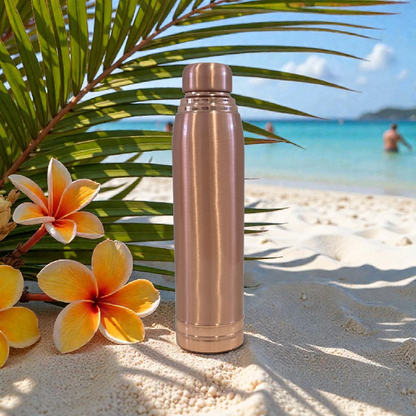 Rose gold water bottle on a sandy beach with flowers and palm leaves